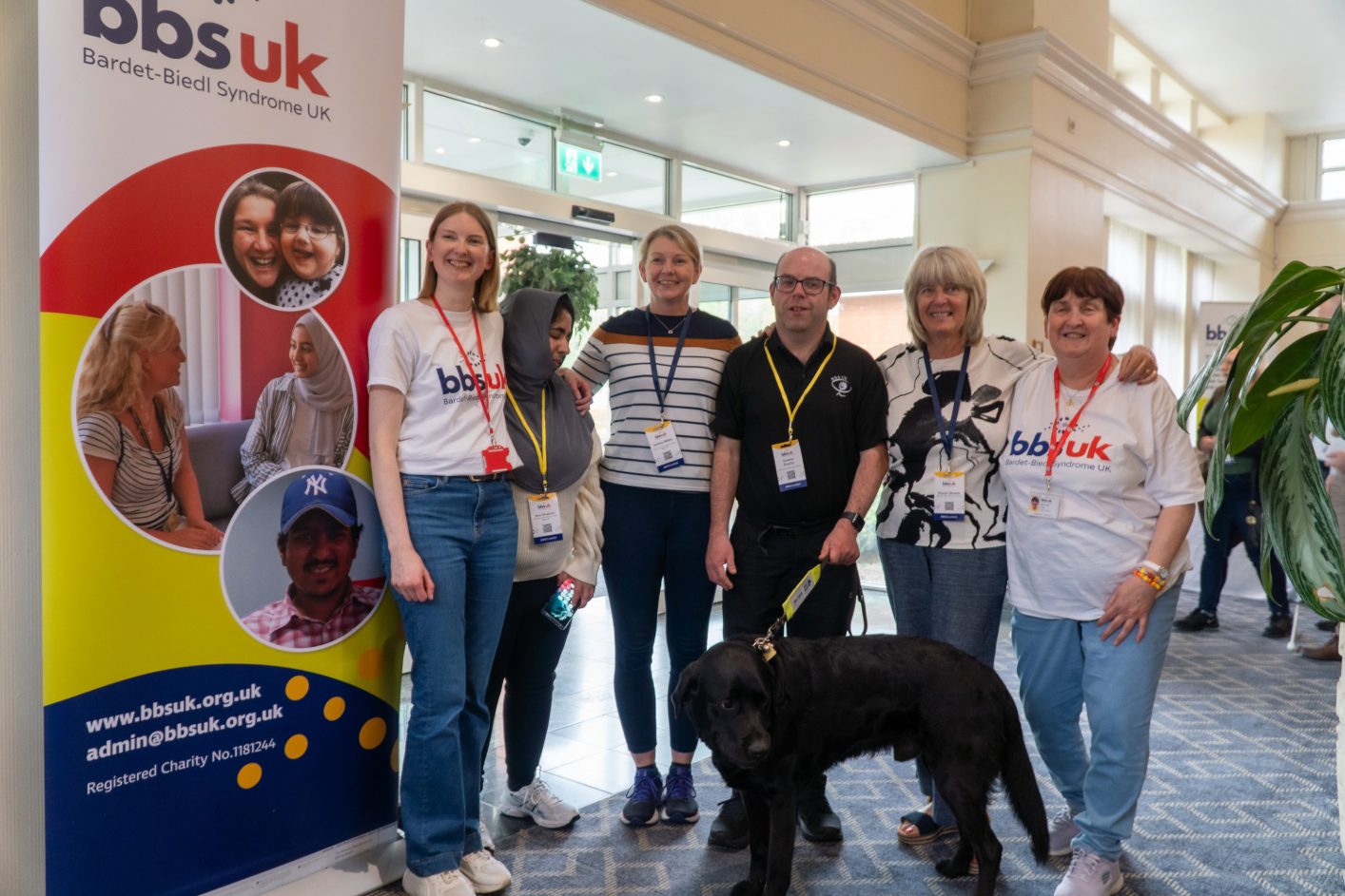 A group of people are stood looking at the camera and smiling. There is a guide dog stood in front of them. There is a BBS UK banner to the side.