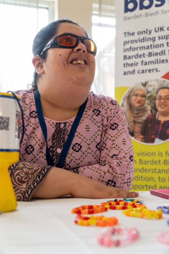 A woman is sat at a table looking up and smiling. She has bracelets in front of her and a collection tin.