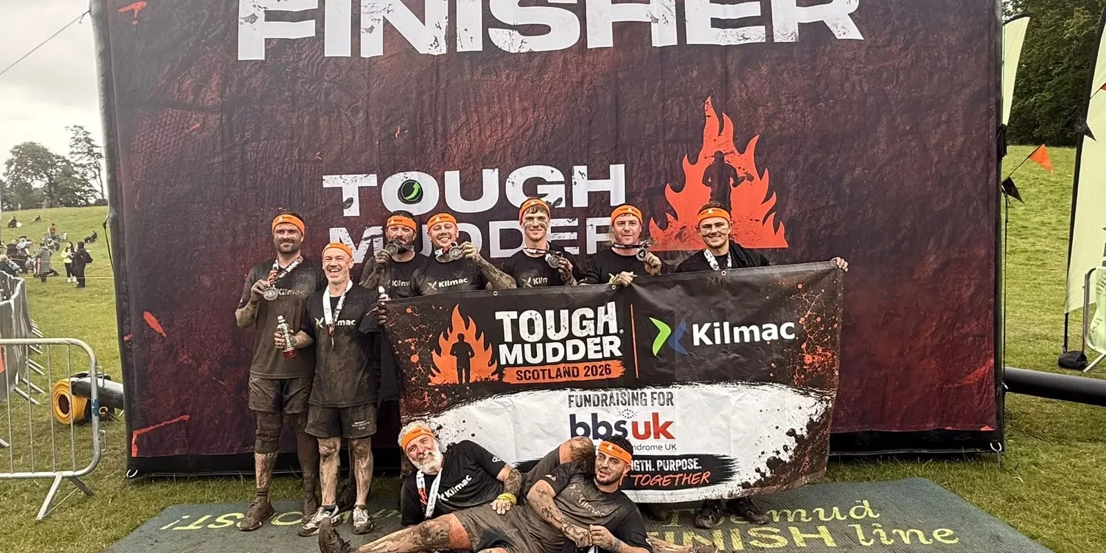 A group of muddy men are holding up a large banner that says Fundraising for BBS UK, shows the Kilmac Ktd logo, as well as the Tough Mudder logo. They are all wearing black tshirts with Kilmac written on the front.