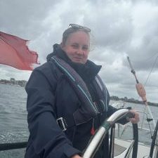 A woman is stood on a boat wearing a life jacket with her hands on the steering wheel.