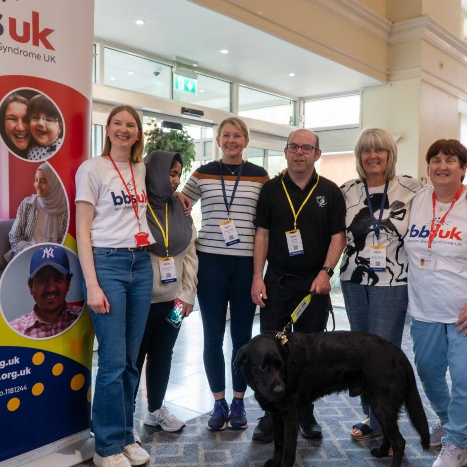 A group of people are stood looking at the camera and smiling. There is a guide dog stood in front of them. There is a BBS UK banner to the side.