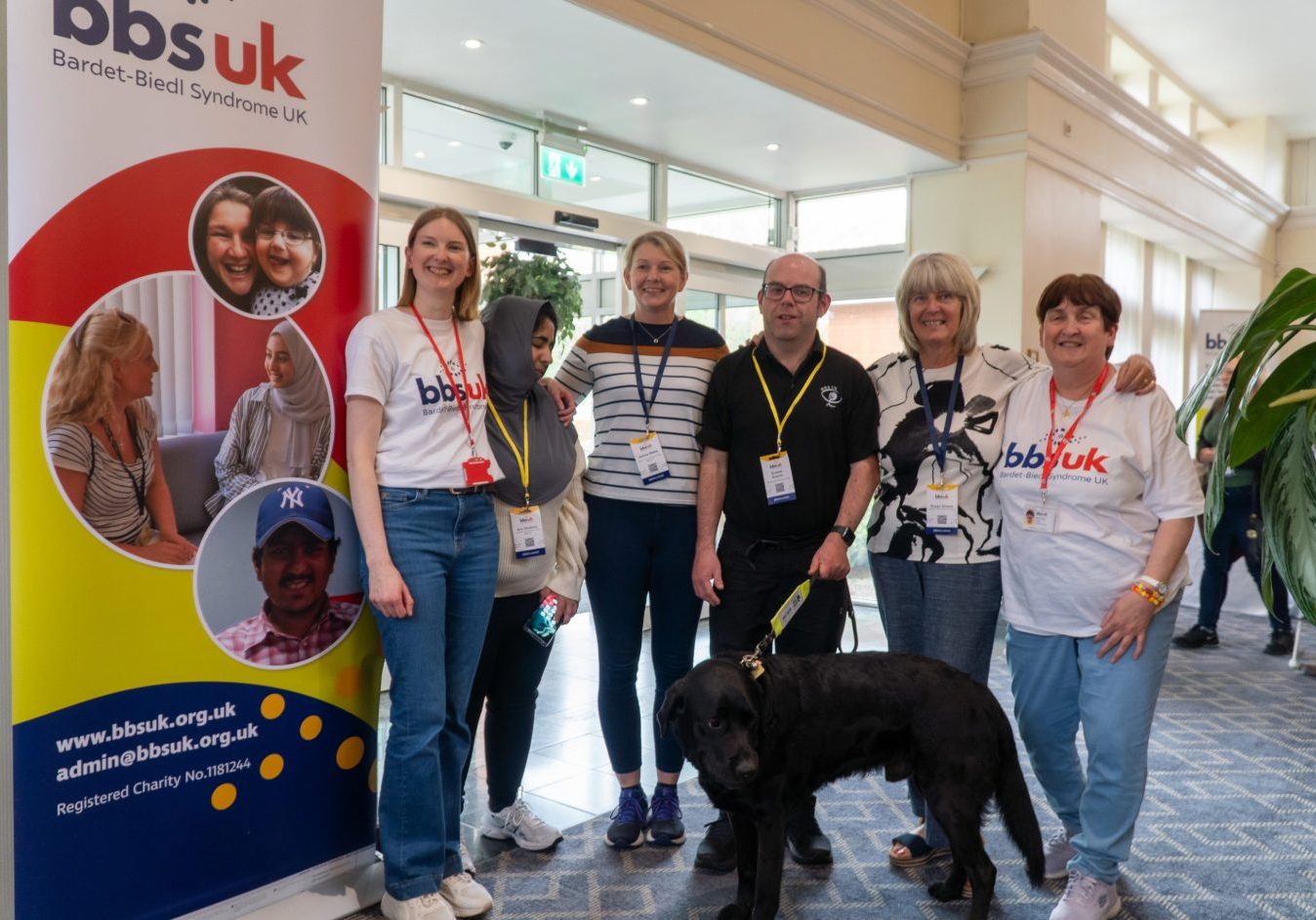 A group of people are stood looking at the camera and smiling. There is a guide dog stood in front of them. There is a BBS UK banner to the side.