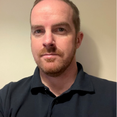 A man is looking at the camera wearing a black polo shirt.