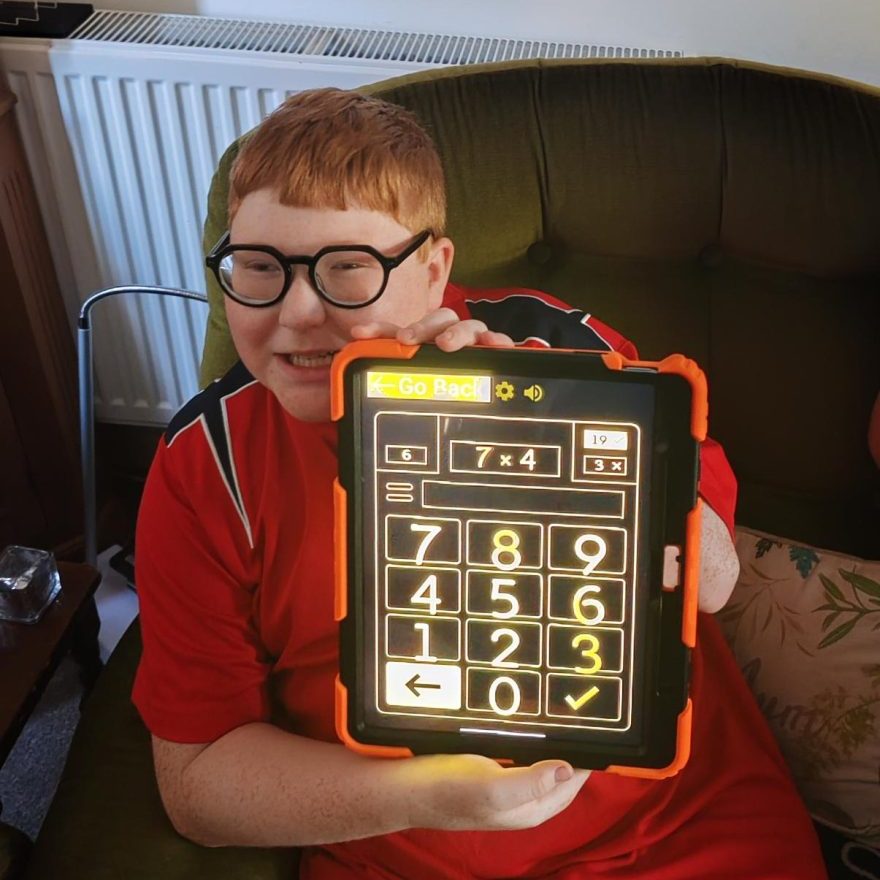 A boy is wearing a red top and holding up a tablet. The display shows a large calculator.