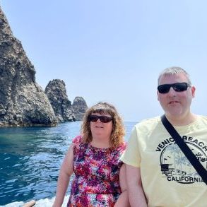 A man and a woman are pictured in front of the ocean, with rocky cliffs behind them. They are both wearing sunglasses. It is a sunny day.