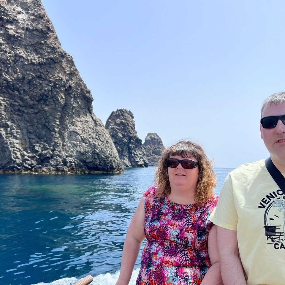 A man and a woman are pictured in front of the ocean, with rocky cliffs behind them. They are both wearing sunglasses. It is a sunny day.