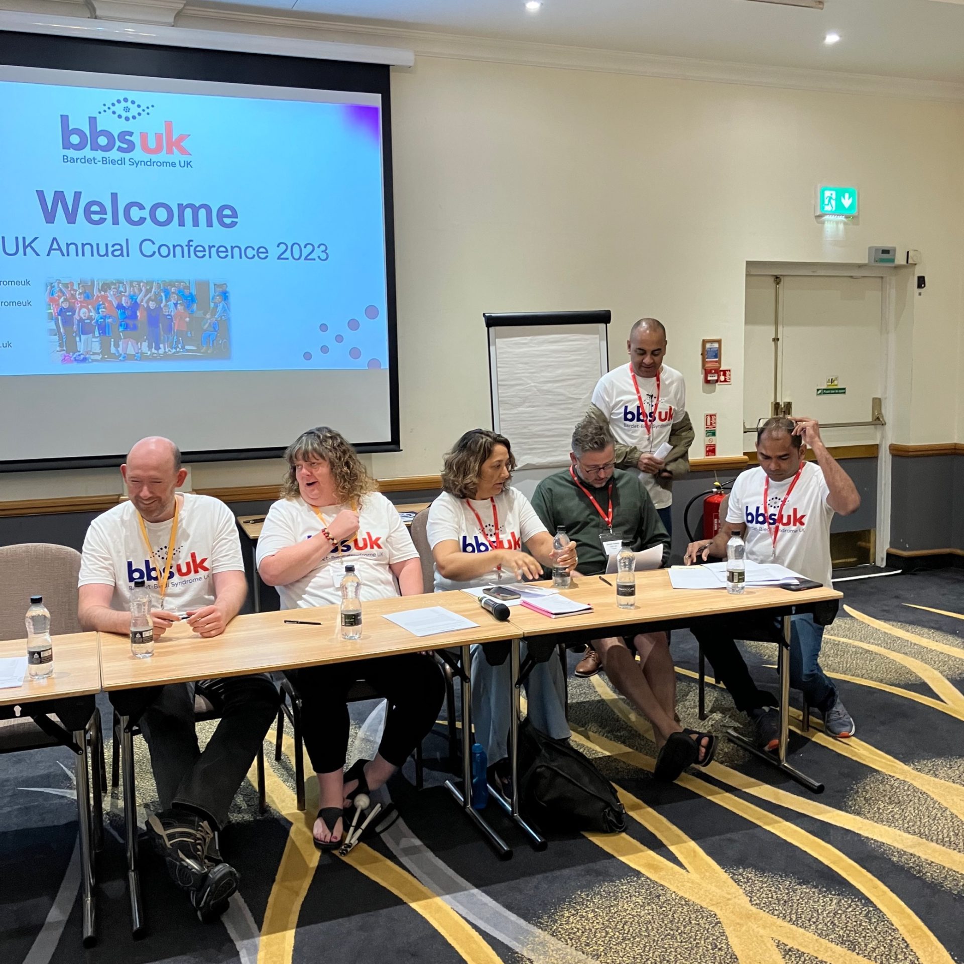 The BBS UK trustees sat at a table with a projector behind them. They are wearing BBS UK t-shirts.